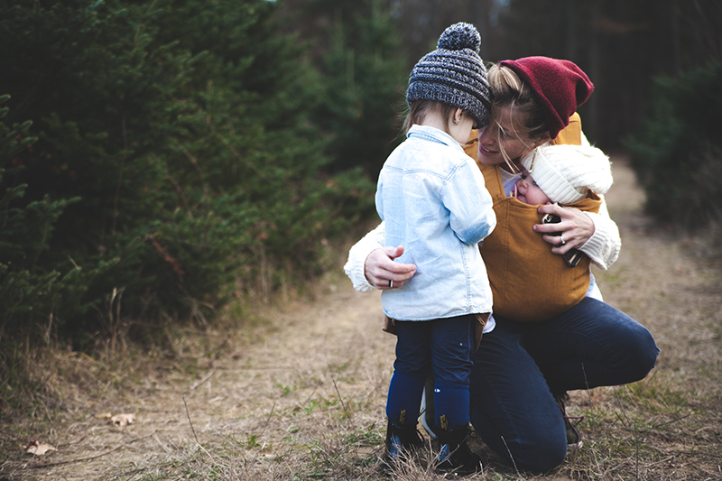 Photo of a woman carrying a baby in a sling, bending now to meet another toddler at eye level for the VancouverMom survey.