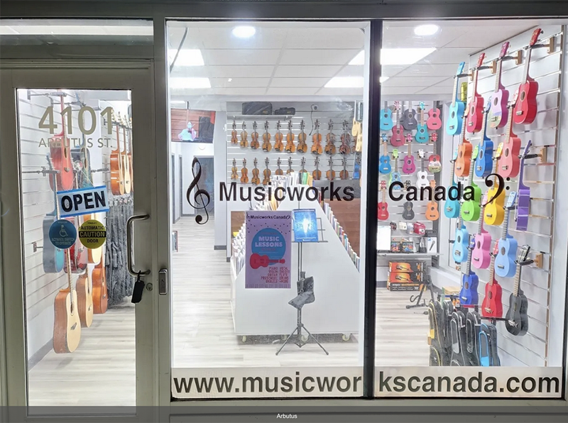 A storefront window for Musicworks. It's brightly lite with rainbow ukulele's hanging on the right. On the left the door sign says open.