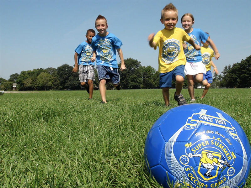 Five little kids in blue and yellow shirts run on green grass after a blue soccer ball that reads Super Summer Soccer Camp.
