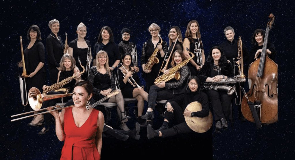 A women in a red dress stands in front of an 18 women orchestra band for Vancouver International Women's Day.