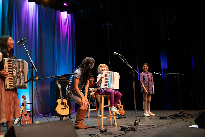 Gina Lina leans in to talk to a little girl sitting on a stool playing the accordian for the 2023 The Children's Festival.