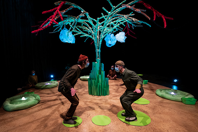 Two men hold their arms out looking at each other. Both are standing on a stage and on top of green pods, a tree is in the background for the 2023 The Children's Festival.