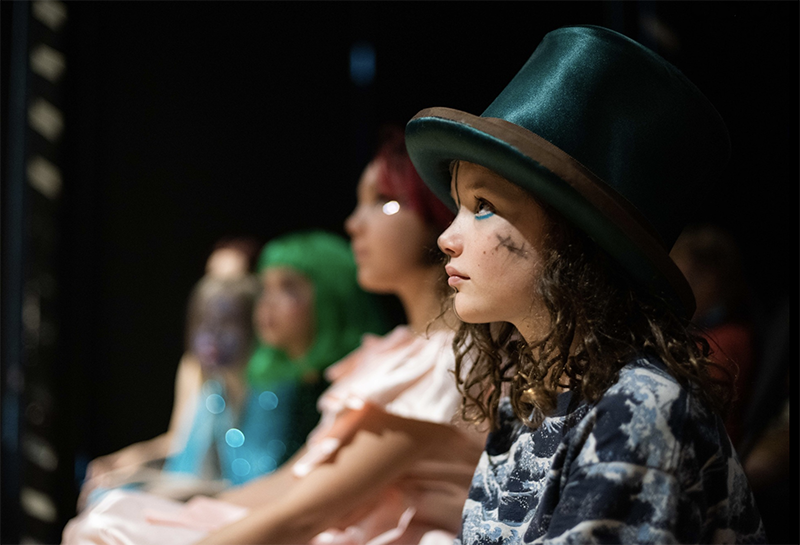 A young girl in a shiny dark green hat and a shirt with ocean waves on it, looks into what we assume is a brightly lit stage.