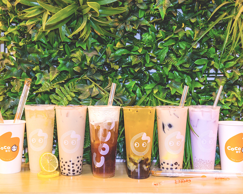 Nine cups of bubble tea sit on a shiny wood table with a leafy green background, all read Coco.