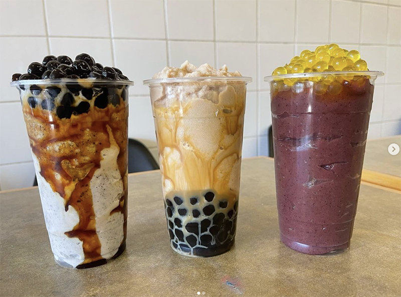 Three bubble teas sit on a table, all three are a slush version of bubble teas. A purple one is topped with fruity pearls.