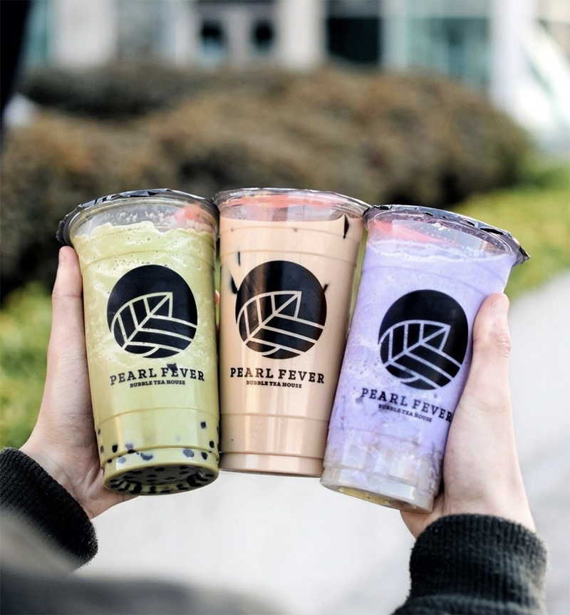 A man holds three bubble teas sandwiched in his hands. The left is green tea, the middle is milk tea and the right is taro, all read Pearl Fever.