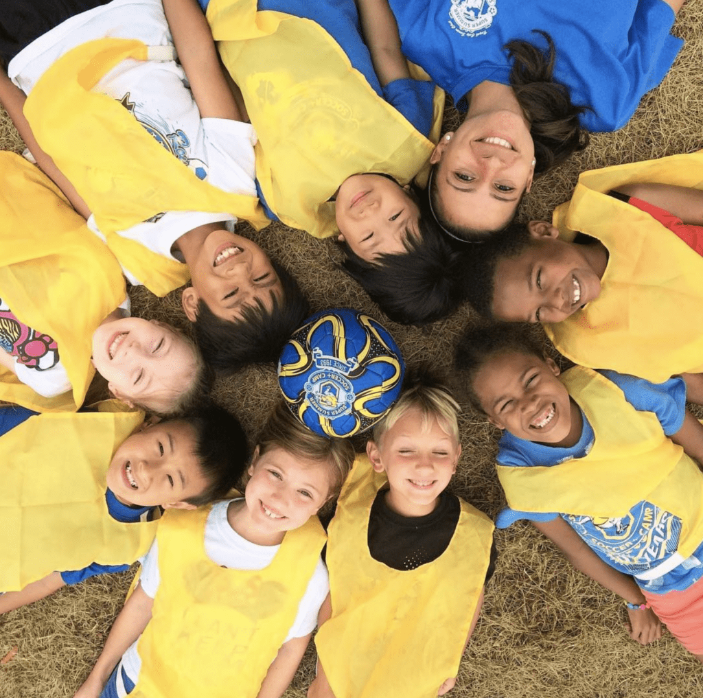 A group of kids laying on the ground in a circle with their heads together with a soccer ball in the middle