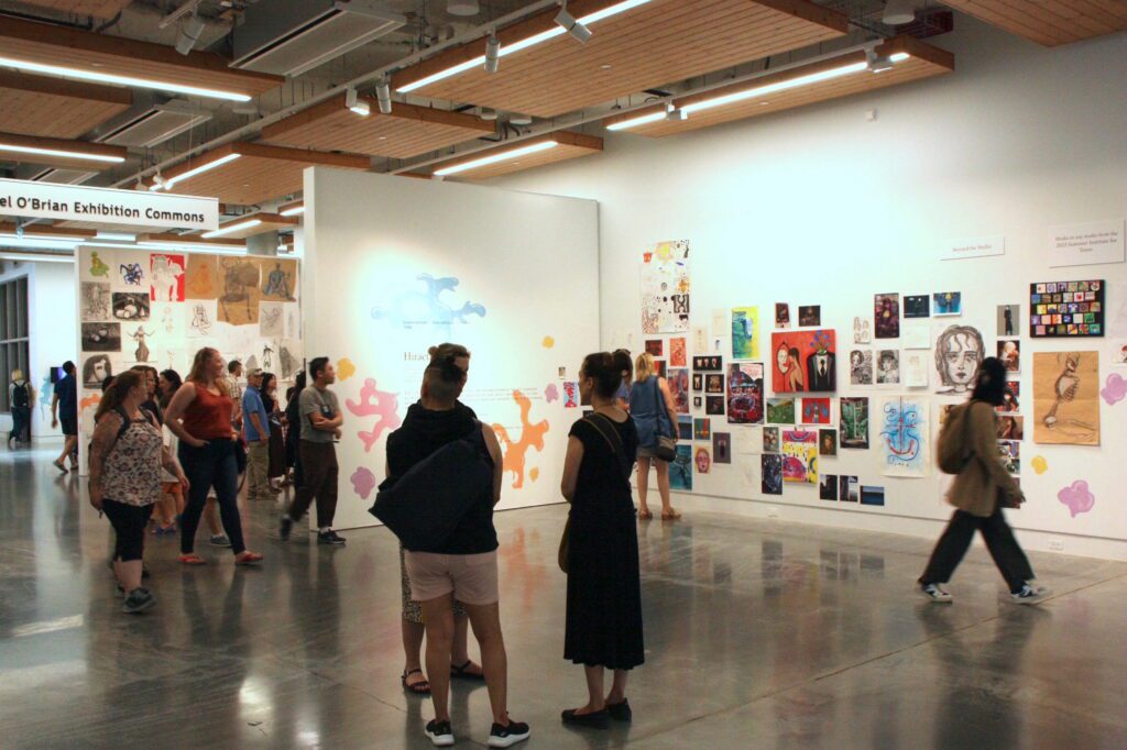 A group of people at the Summer Teen Program Exhibition for the Emily Carr University students. You see a showcase of artwork displayed on the wall.