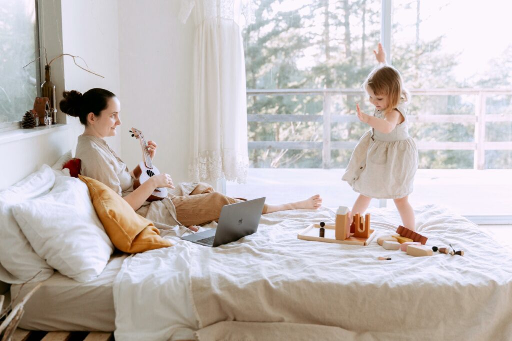 Mom sitting on her bed playing the ukelele and her daughter dancing on the bed as these are indoor activities to do while snowing