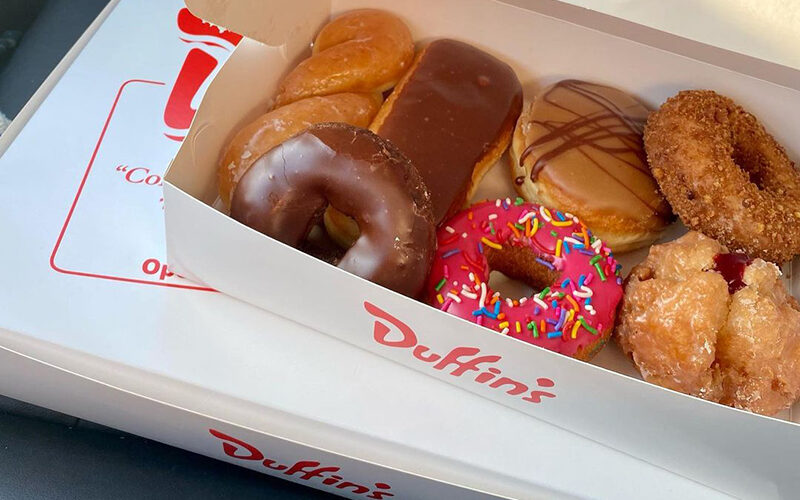 An open box of seven donuts sits on another closed box. There is a twist, a longjohn, a maple filled, a cinnamon, a chocolate dip, a rainbow sprinkle, and a raspberry cream filled donuts.